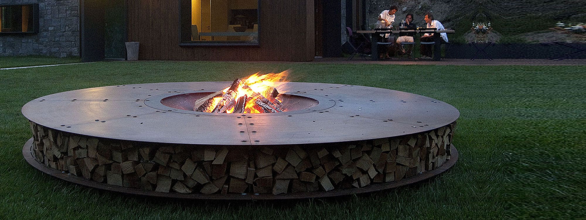 Image of garden at dusk with flames dancing within AK47 Zero large circular fire pit's burning chamber