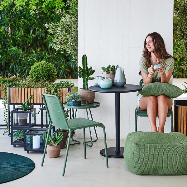 Image of woman sat on olive-green Copenhagen chair, resting her feet on Divine crocheted foot rest, surrounded by Combine modern planters by Cane-line