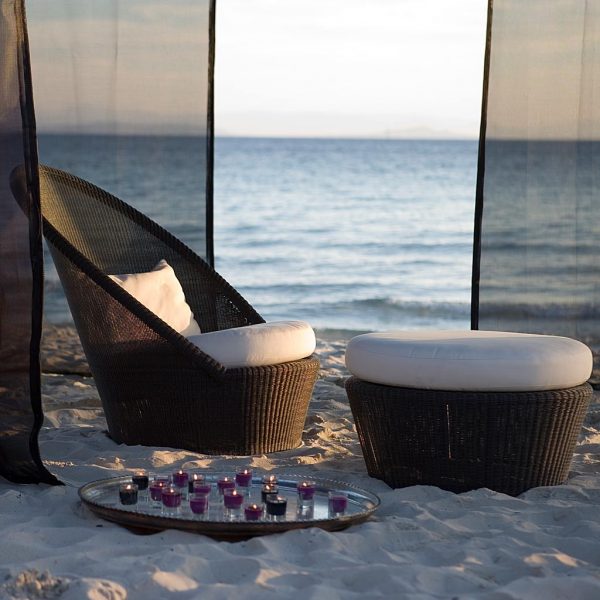 Image of mocca-coloured Kingston garden easy chair and footstool by Caneline, shown in the sand on a beach with drapes and sea in background