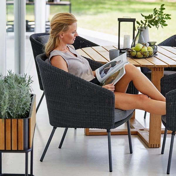 Image of woman sat reading magazine in Mega garden chair next to Endless circular teak table by Cane-line