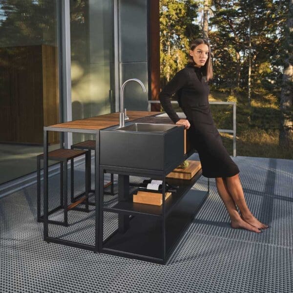 Woman seated at Open Bistro outdoor kitchen furniture beside Open Kitchen BBQ and sink by Roshults on a minimalist garden terrace