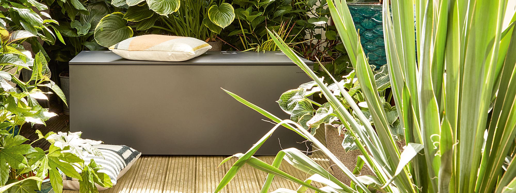 Image of El Pecho anthracite outdoor cushion box by Conmoto, shown on wooden decking surrounded by exotic plants