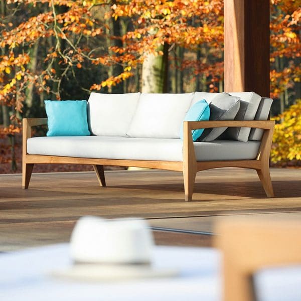 Image of Royal Botania Zenhit Teak Garden Sofa with grey and blue cushions with Autumnal trees in background