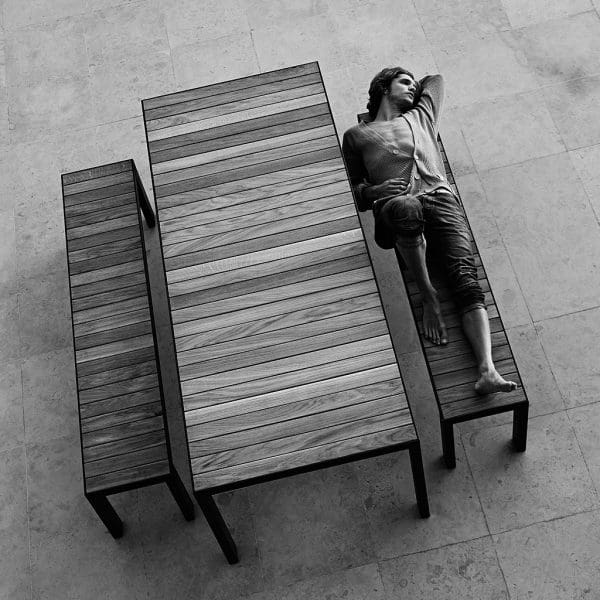Monochrome image of dude in cardigan lying on Roshults modern garden bench in teak & anthracite stainless steel