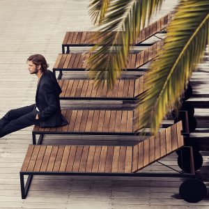 Image of dude in black suit sitting n end of Roshults teak and stainless steel sun lounger
