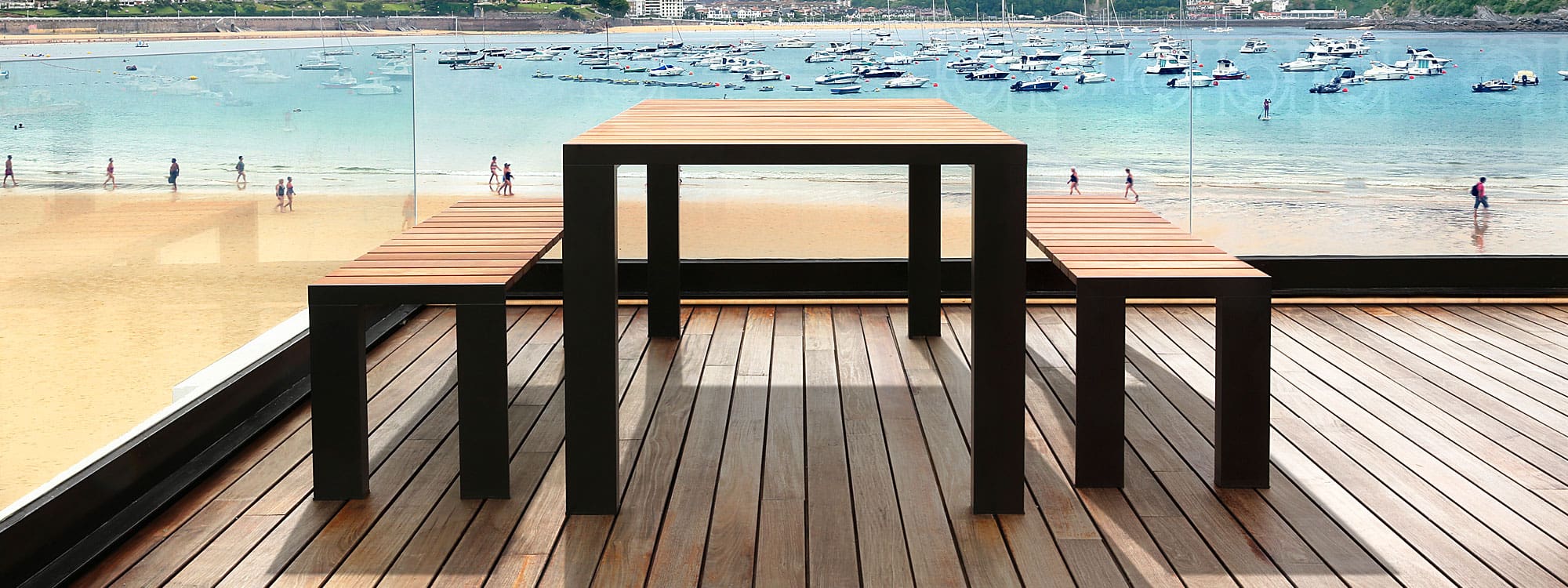Image of Stua Deneb modern garden table and benches with black aluminium frames and teak slats, shown in wooden decked terrace with multiple moored boats in a bay in the background