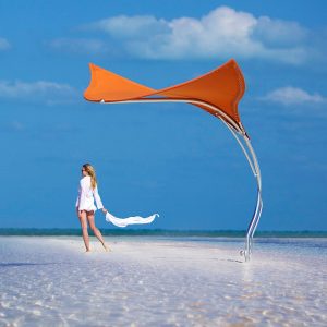Image of woman walking along a tropical sandy beach next to orange Tuuci Stingray parasol with electro-polished stainless steel mast, with blue sky in the background