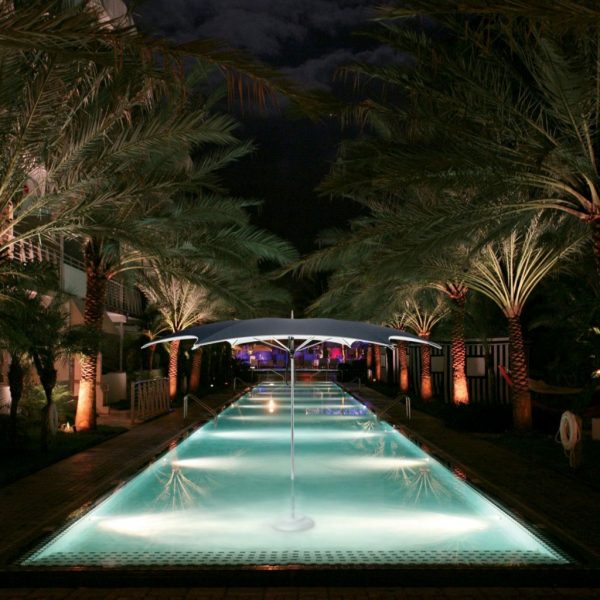 Nighttime image of Ocean Master Max Crescent center pole parasol mounted in illuminated shallow water of swimming pool