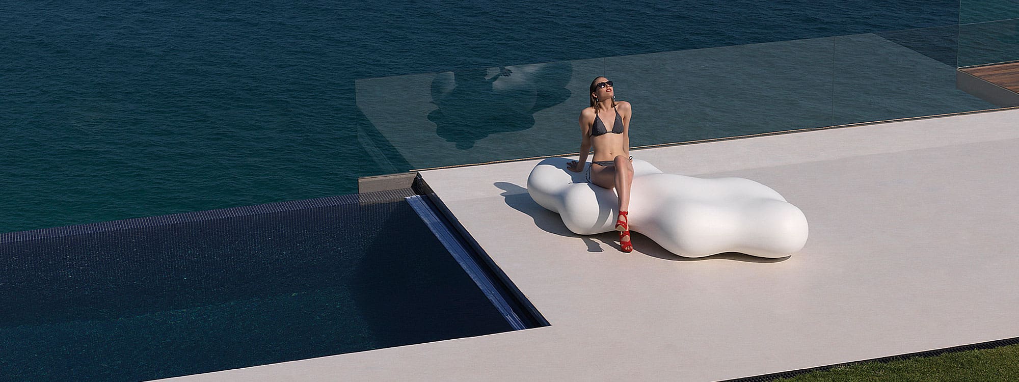 Image of woman sat in sunshine on Vondom Lava amorphous outdoor bench, shown on poolside