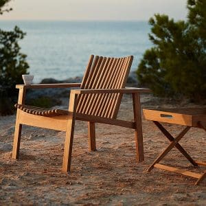 Image of Cane-line Amaze teak lounge chair on earth, with pine trees and sea in the background