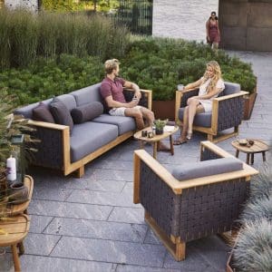 Image of man and woman sat chatting on Cane-line Angle teak sofa and lounge chair, with Grey Airtouch cushions and grey SoftRope webbing