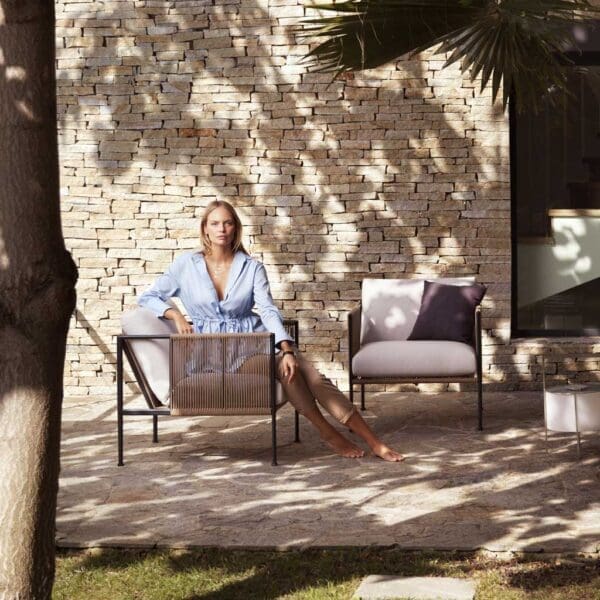 Image of woman sat on Antibes 2 seat outdoor sofa in shady courtyard next to palm tree