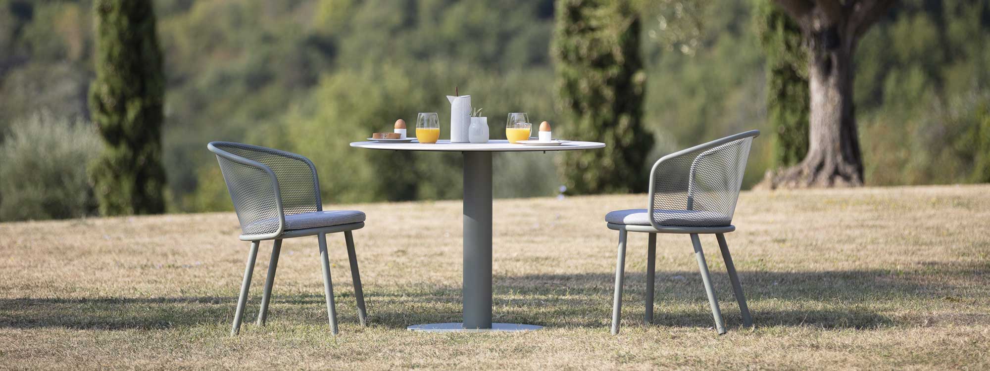 Image of taupe Baza modern garden chairs and Branta small round garden table on scorched grass with trees in the background
