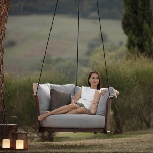 Image of woman sat relaxing in Baza modern garden swing seat beneath tree, with Luci garden lanterns to one side, and arid countryside in background