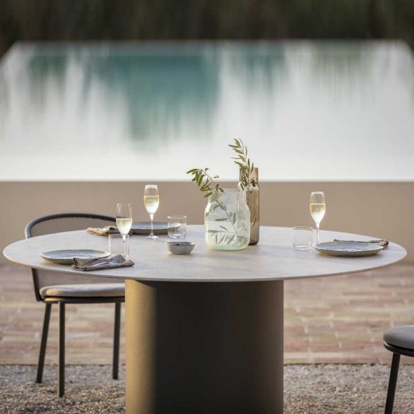 Image of Branta modern round garden table with glasses of white wine and vase of olive leaves set on table top, with tranquil swimming pool in background in soft light of dusk