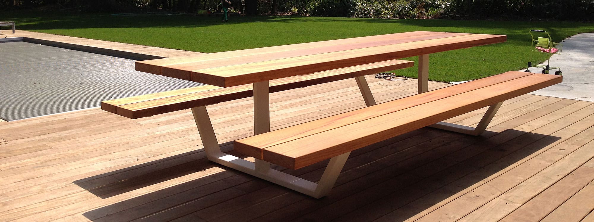 Image of Cassecroute minimalist picnic table and benches with white frame and iroko hardwood surfaces, shown on wooden decking next to swimming pool