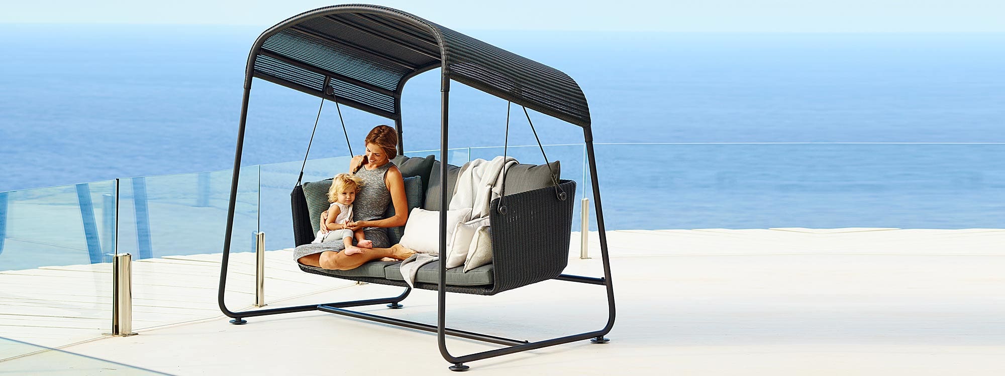 Image of woman and child sat in Cane-line Cave black swing seat with grey cushions on sleek terrace with sea in background disappearing into horizon