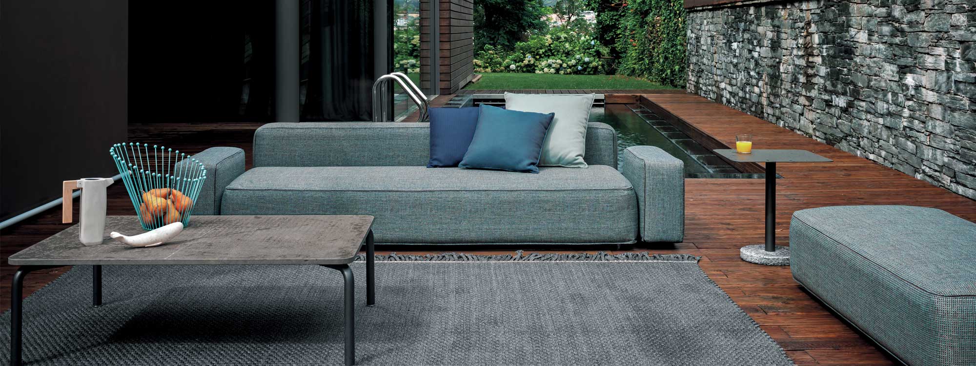 Image of Dandy minimalist grey garden sofa and ottoman, together with Piper stone topped coffee table and Bernardo side table, with chic plunge pool and garden in the background