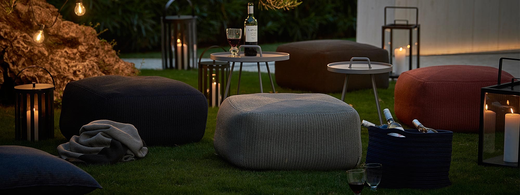 Nighttime image of Cane-line Divine crocheted garden poufs together with On The Move outdoor tray tables and Lighthouse lanterns
