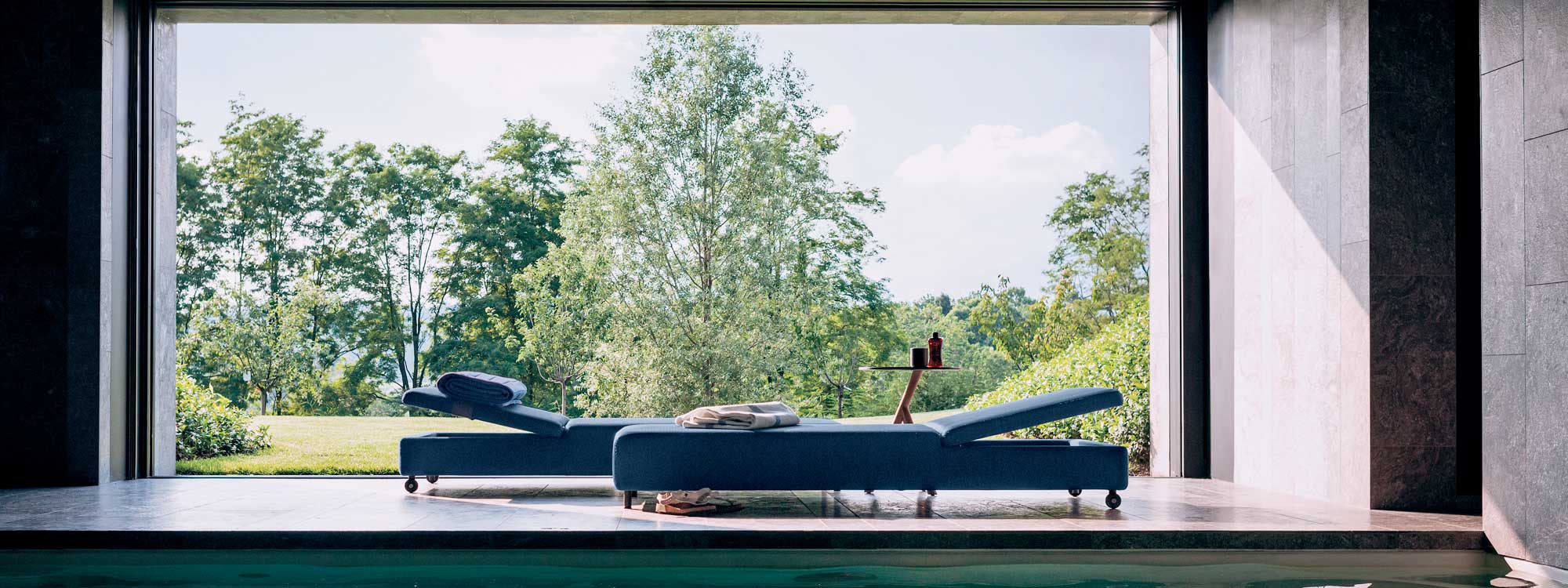 Image of pair of Double upholstered sun loungers by Rodolfo Dordini for RODA, shown on minimalist indoor poolside with garden and trees in the background