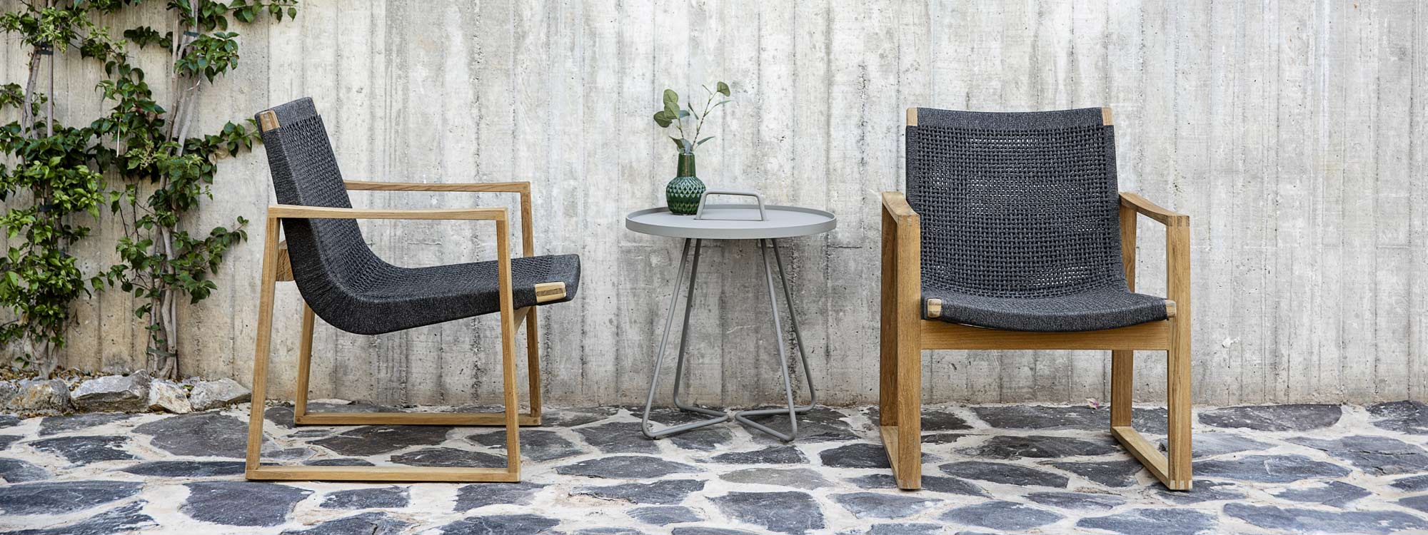 Image of pair of Cane-line Endless lounge chairs and taupe On The Move tray table on stone terrace against concrete wall with shutter boarding indentations