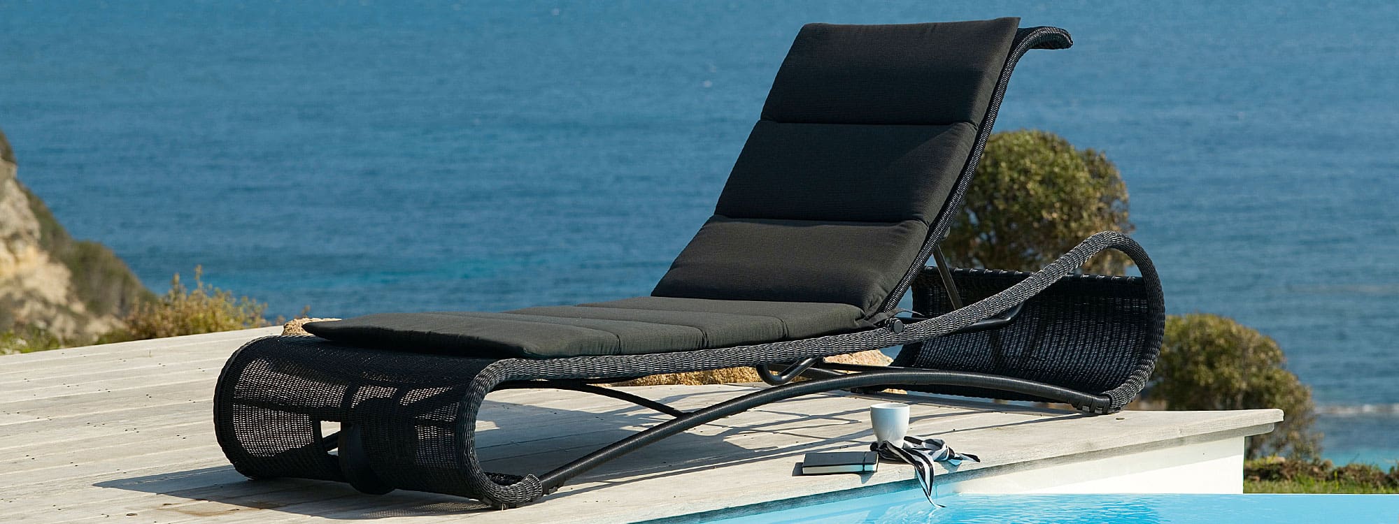 Image of black rattan Escape sun lounger with black cushion by Cane-line, on poolside with sea in the background