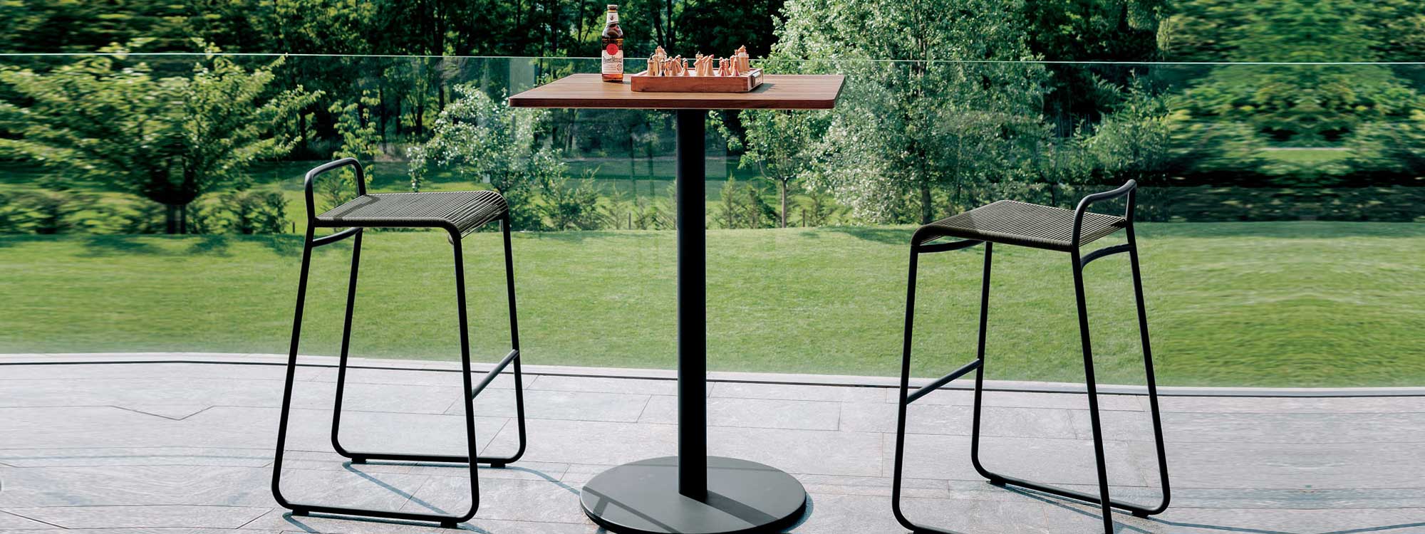 Image of pair of RODA Harp bar stools with tubular frame and acrylic cord seat and back, together with Stem pedestal bar table on terrace, with garden in background