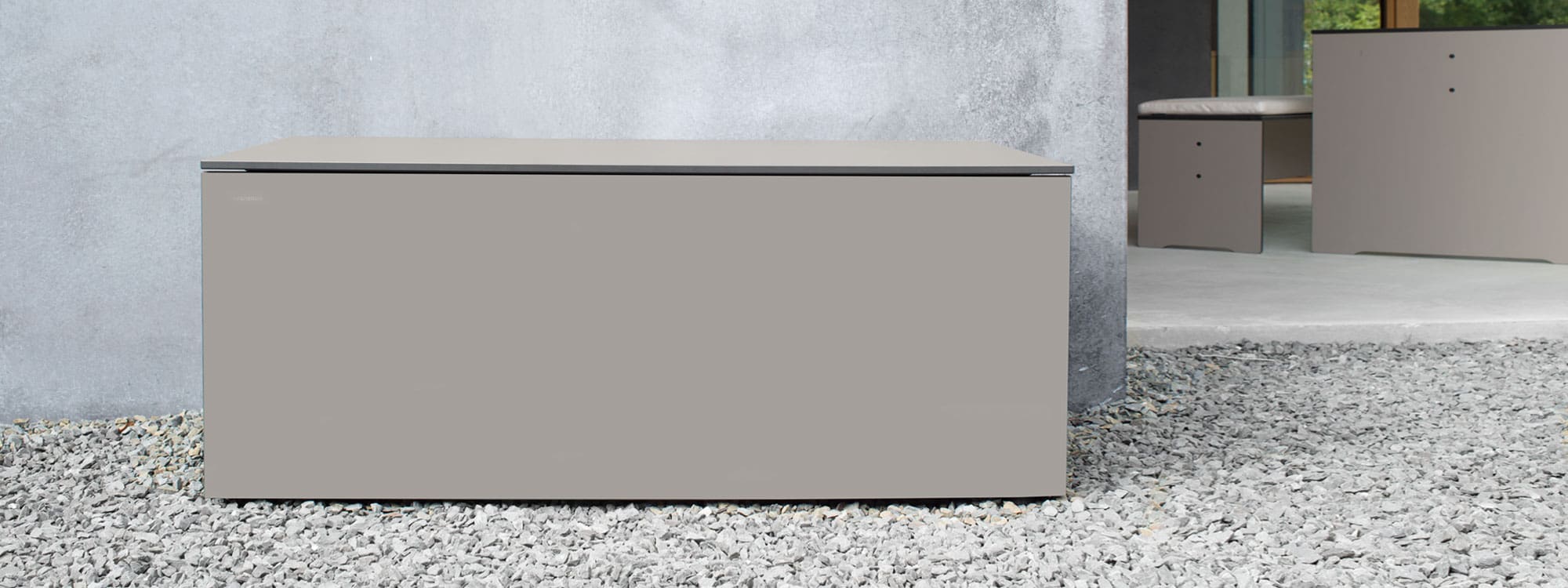 Image of Conmoto El Pecho taupe coloured garden storage box, shown on gravel, with poured concrete wall and Riva HPL furniture in the background