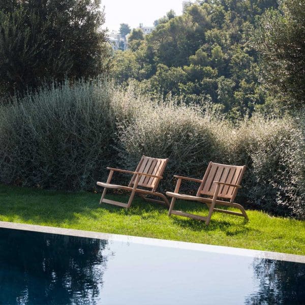 Image of 2 RODA Levante teak garden lounge chairs on lawn beside horizon pool with reflection of trees in the water