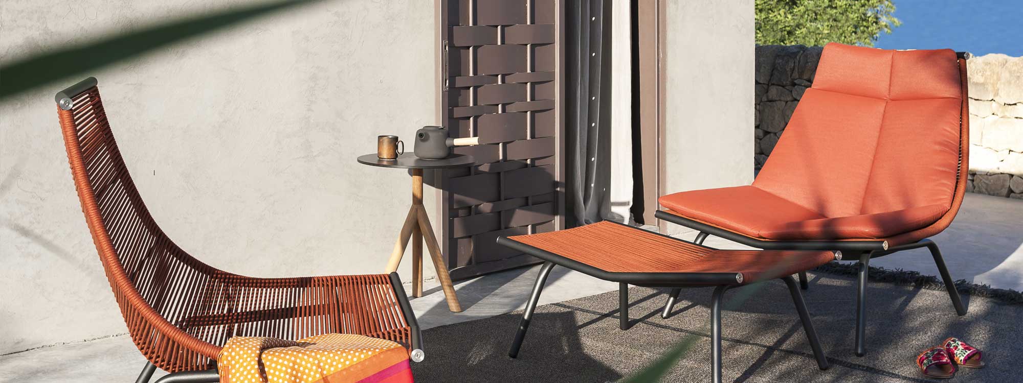 Image of pair of RODA Laze minimalist outdoor lounge chairs with smoke-colored frame and orange cord seat and back, together with Laze foot rest in the same finish