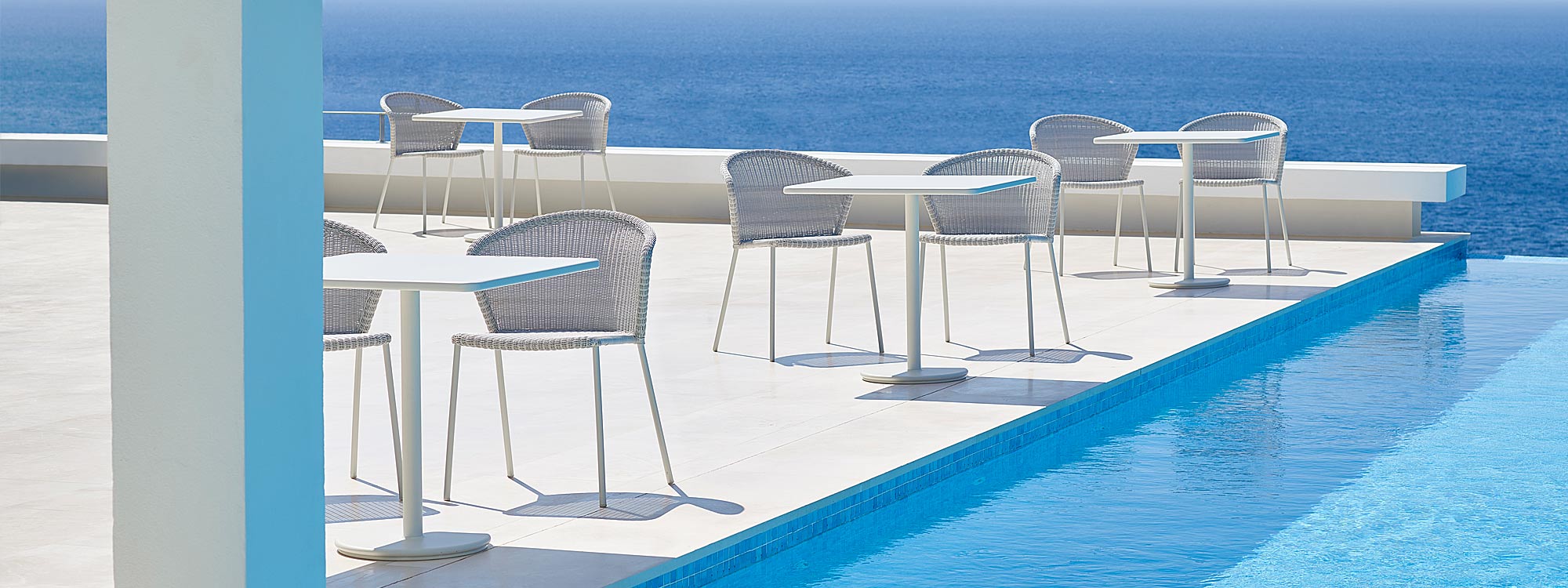 Image of white-grey Cane-line Lean chairs and white Go bistro tables on sunny poolside with sea in background