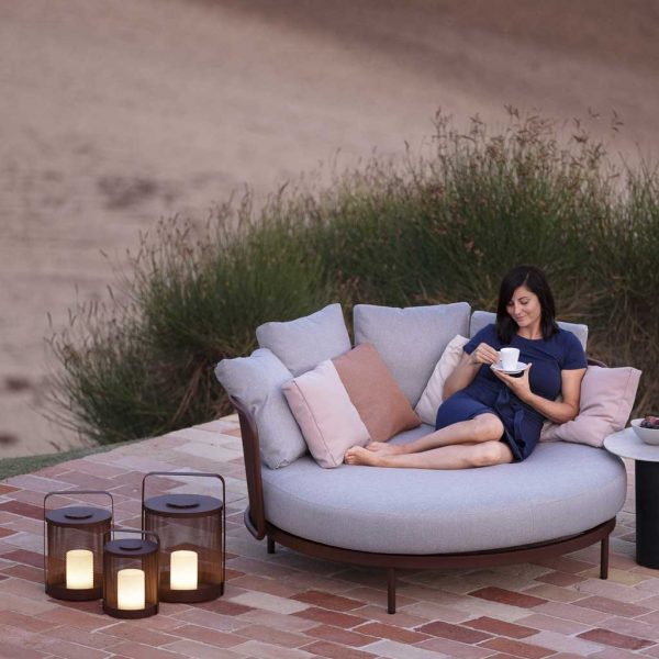 Image of woman relaxing on Baza daybed next to 3 Rust-Brown coloured Luci LED garden lanterns