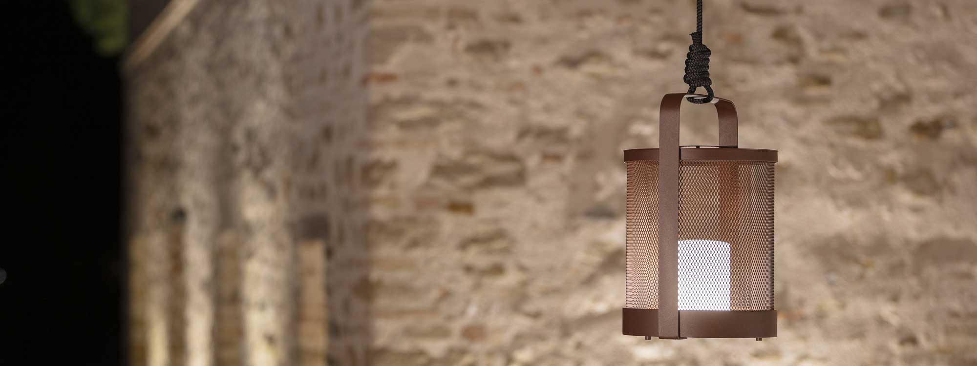 Image of Luci outdoor LED lantern in Rust-Brown suspended from rope with rustic stone wall in background