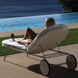 Image of woman lying on Shell modern stainless steel lounger by FueraDentro, on sunny poolside overlooking horizon swimming pool and the sea in the distance