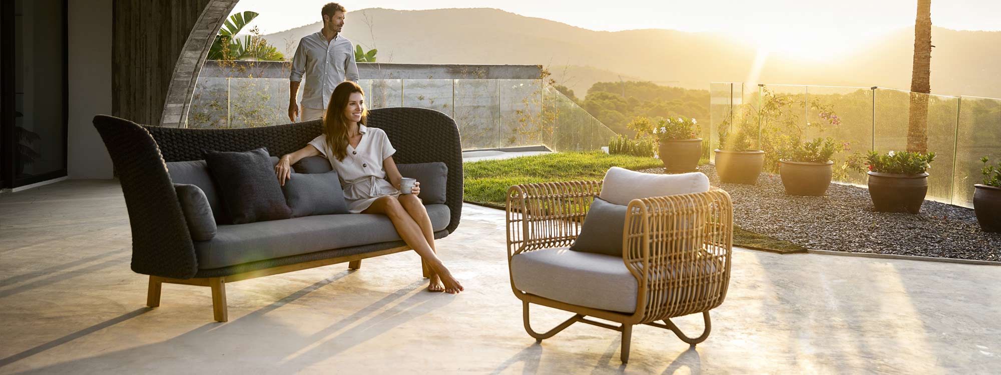 Image of sunset terrace with woman sat on Caneline Peacock Wing dark grey garden sofa next to Nest synthetic cane lounge chair