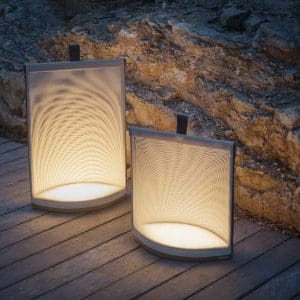 Image of pair of RODA Pillow minimalist outdoor electric lanterns on decking, giving off soft white light against a drystone wall