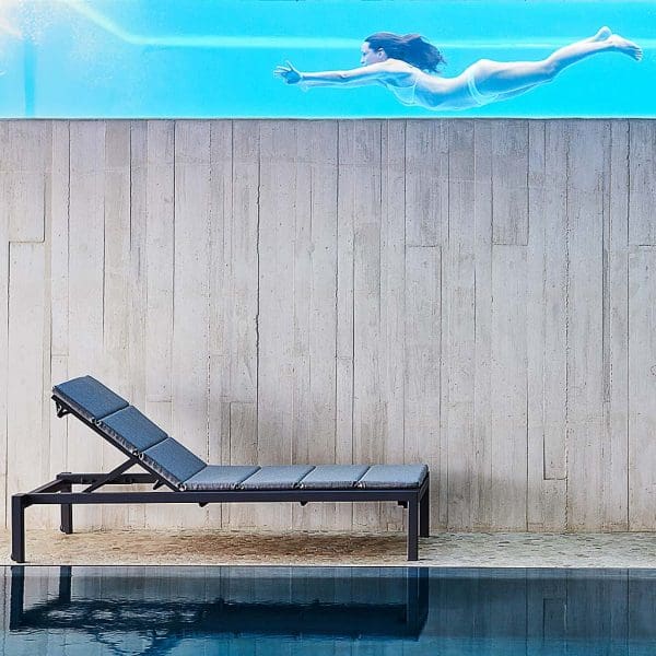 Image of Dark-grey Relax sun lounger with dark-grey cushion by Cane-line, shown on poolside with woman swimming in another pool above