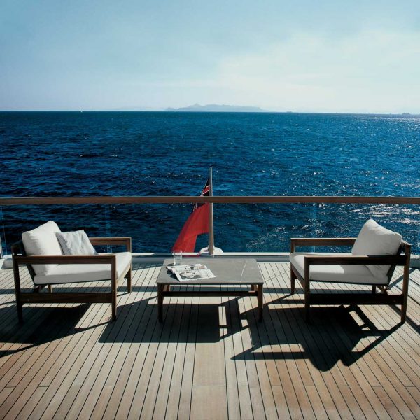Image of pair of RODA Road modern teak lounge chairs and low table on aft deck of super yacht with fluttering ensign flag and sea in the background