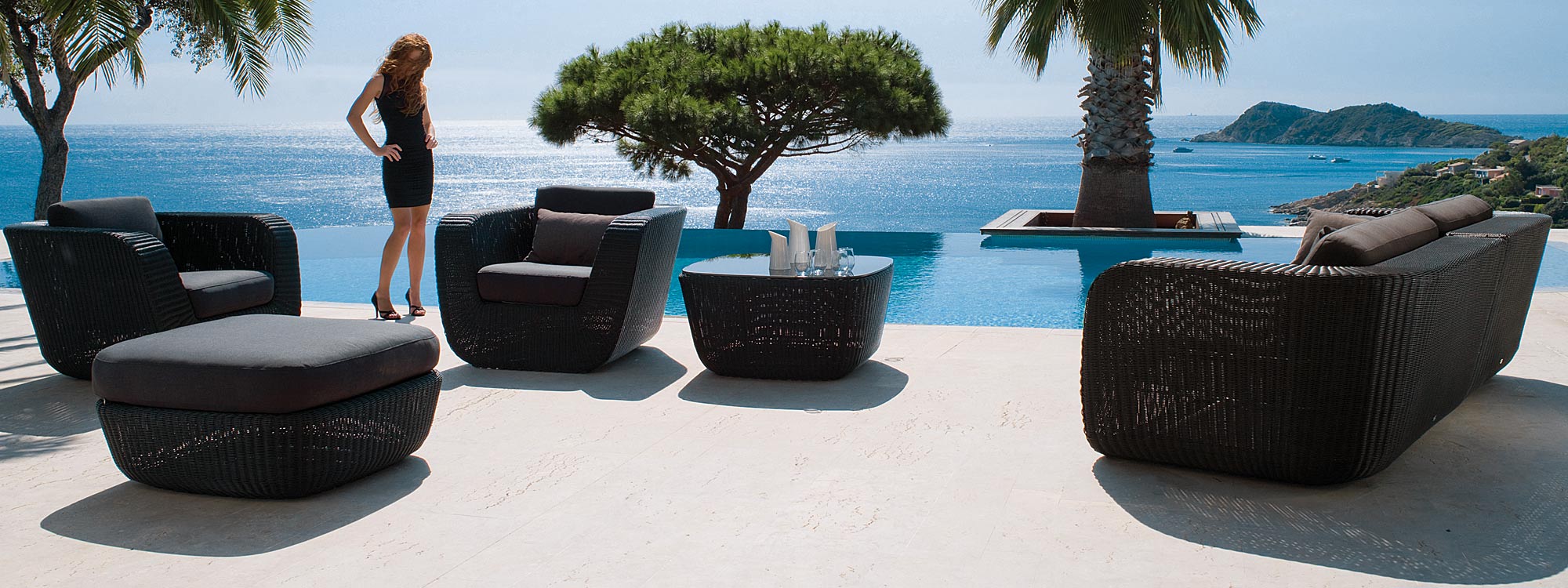 Image of woman in black dress next to Savannah black garden sofa set by Cane-line, with sparkling sea in the background