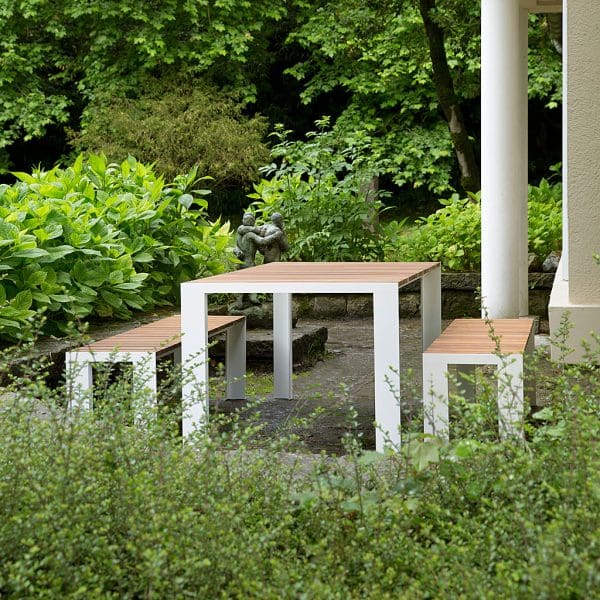 Image of Stua Deneb minimalist outdoor table and benches with white aluminium frames and slatted teak surfaces, on terrace surrounded by shrubs
