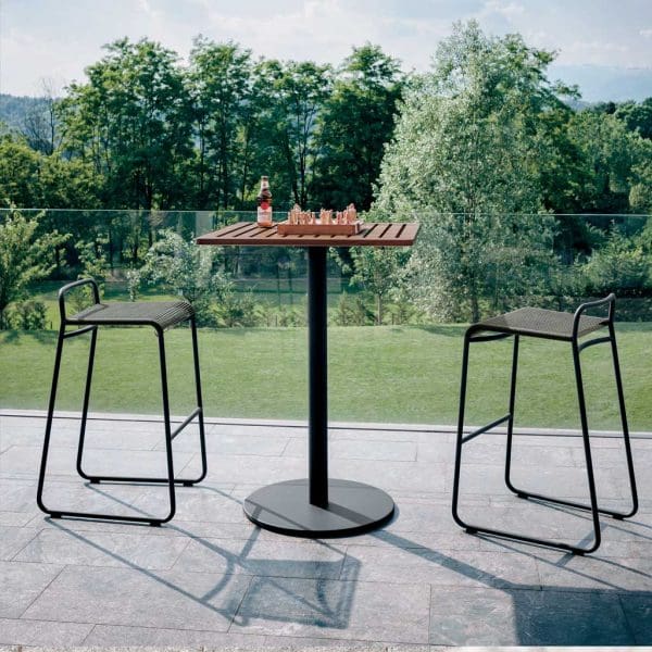 Image of RODA Button tall bistro table with Harp modern garden bar chairs, shown on terrace with lawn and shrubs in background