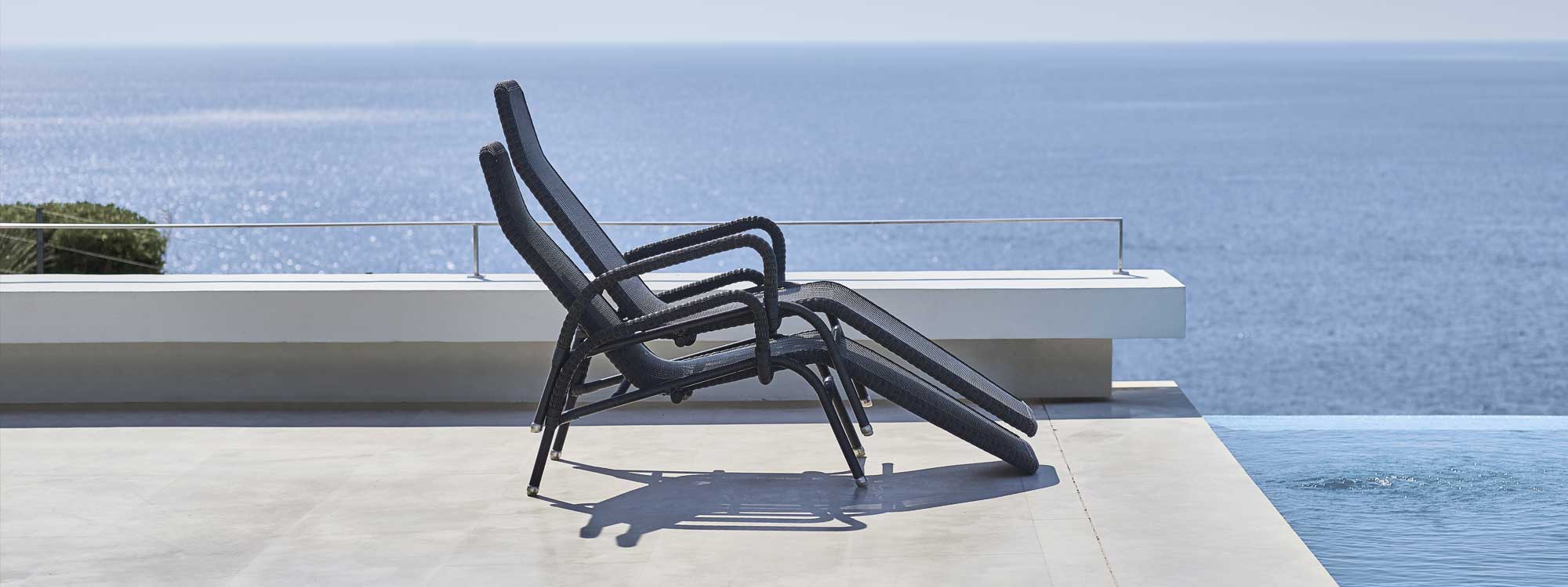 Image of pair of stacked Sunrise recliner chairs by Cane-line, shown next to swimming pool with sea in the background