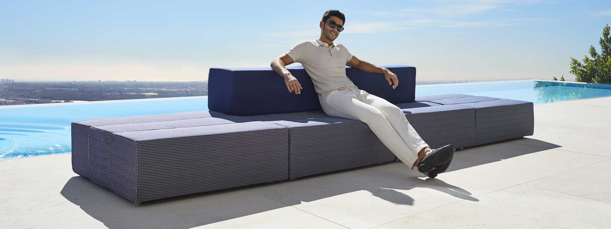 Image of young dude with sunglasses sat on Vondom Tablet blue garden sofa, with horizon swimming pool and blue sky in the background