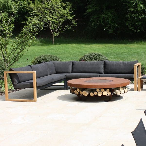 Image of FueraDentro Cima Lounge modern teak corner sofa with grey cushions, with Ercole fire pit in the foreground and lawn and hedge in the background