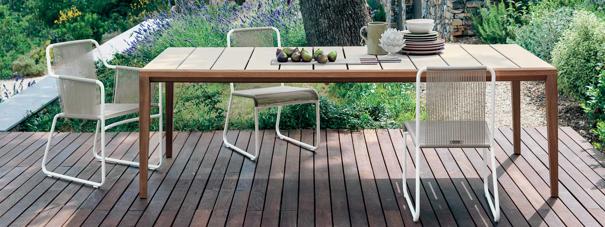 Image of RODA Teka teak table with table top in slats of mat ceramic, with Harp white garden chairs with seat & back in brown cord, with trees and lavender in background