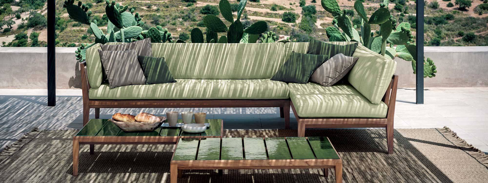 Image of RODA Teka teak corner sofa with light green cushions beneath pergola, with cactuses and arid countryside in background
