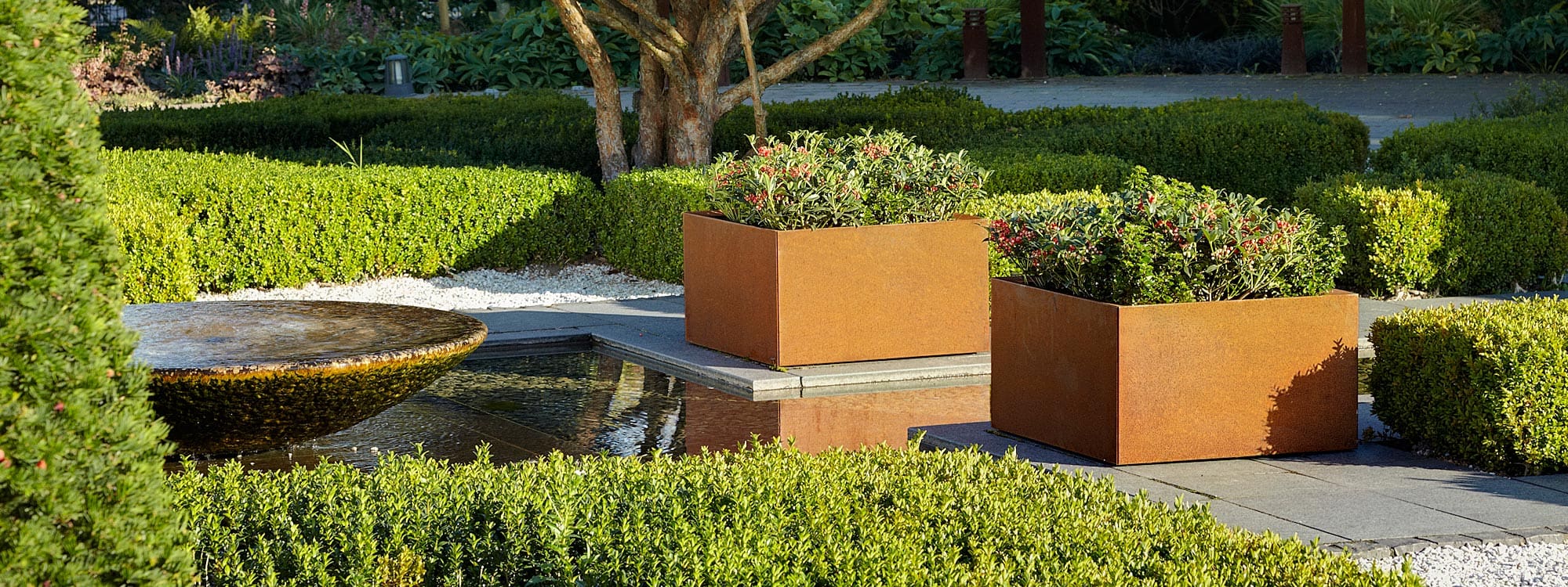 Image of pair of Thallo oxidised corten planters by Flora, shown next to still waters within a parterre garden
