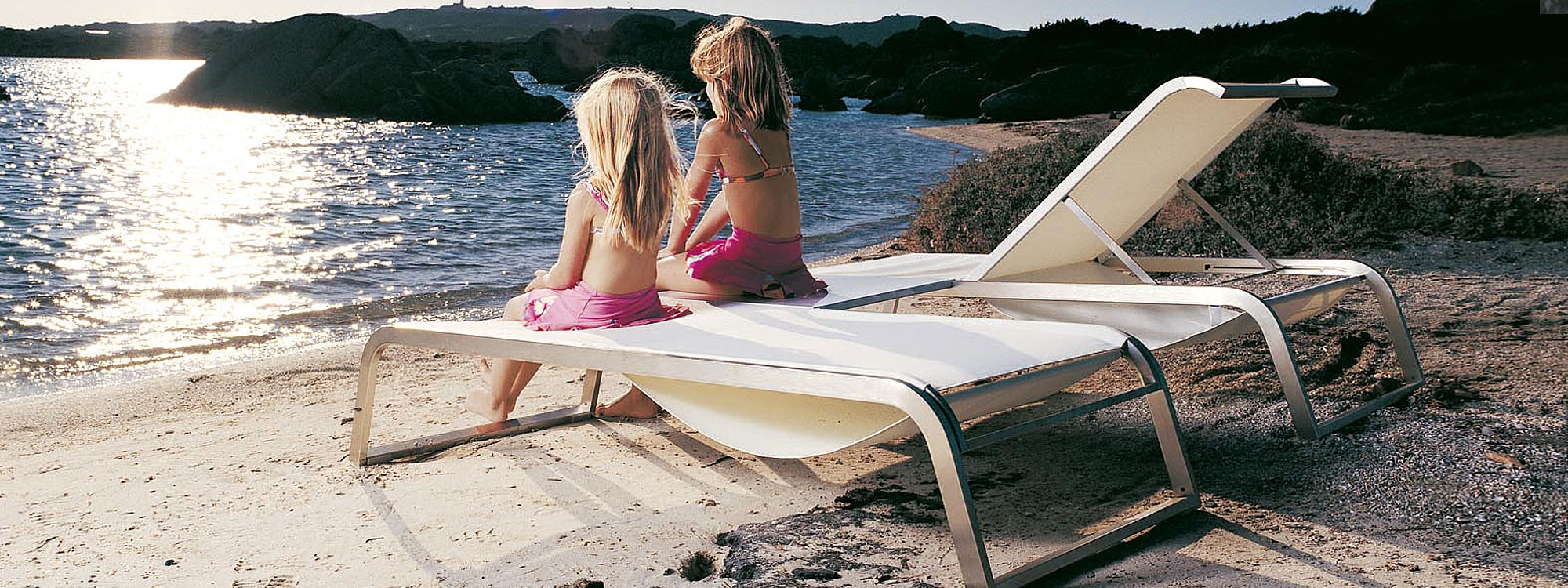 Image of pair of young girls sat on Coro L3 white sun loungers on sandy beach with sunlight glinting on the water