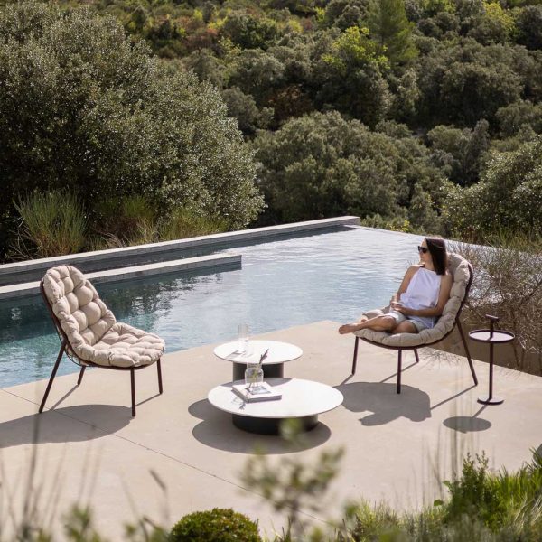 Image of woman sat with her feet up on Albus garden easy chair, with Branta low table in the centre, with swimming pool and woodland in background
