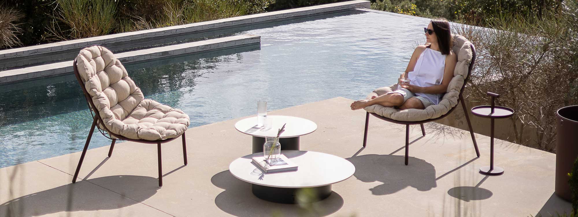 Image of woman sat with her feet up on high-back Albus garden lounge chair on sunny poolside, with Branta low tables and another Albus relax chair to one side